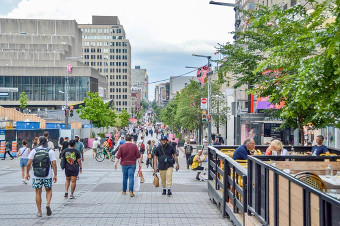 People walk, sit, and dine along a pedestrianized street in Montreal. It is lined with trees, planter boxes, terraces, and benches.