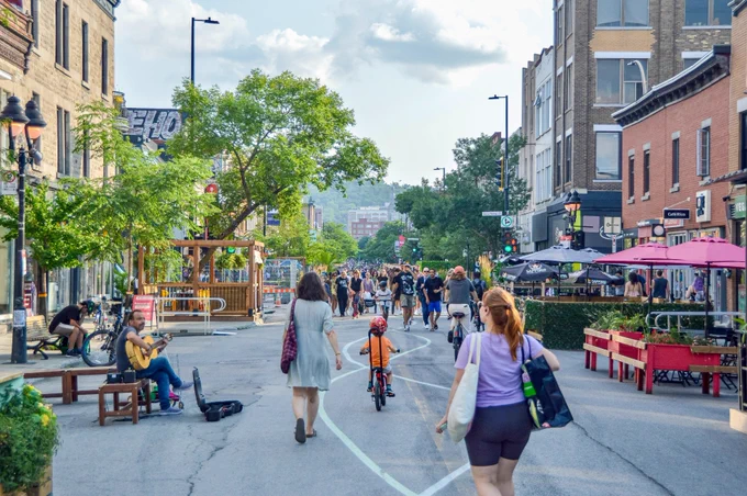 People walk, sit, and dine along a pedestrianized street in Montreal. It is lined with trees, planter boxes, terraces, and benches.