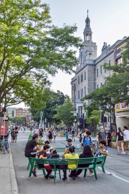 People walk, sit, and dine along a pedestrianized street in Montreal. It is lined with trees, planter boxes, terraces, and benches.