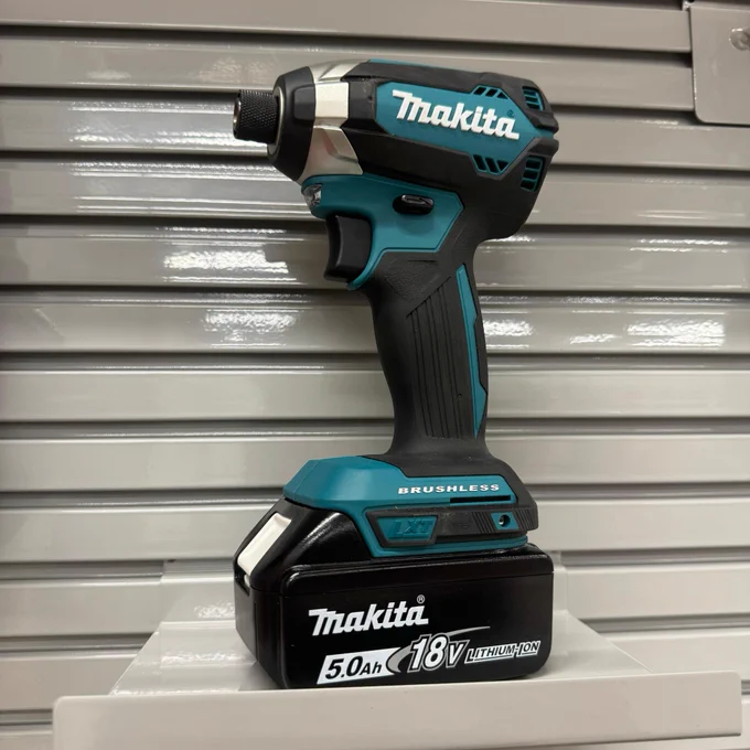 An upright Makita brand brushless power drill displayed on a shelf. The drill is colored in teal and black, with the Makita logo prominently displayed in white on the side. It features an 18V, 5.0Ah battery attached at the base. The background consists of horizontal grey metal slats.
