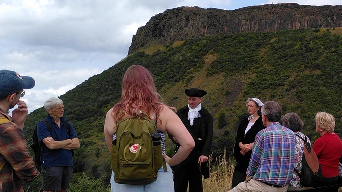 Actors Billy Quain as James Hutton and Jane Westhead playing Isabella, James' sister, entertain a crowd in Holyrood Park with Salisbury Crags in the background.