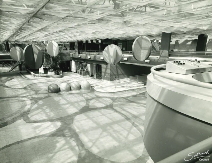 Black and white photograph showing a wide view of a swimming pool. There are oval patterns on the bottom of the pool and hanging spherical decorations from the ceiling. The ceiling appears to be made from perspex panels in pyramid shapes and natural light is flooding the swimming pool. There is a control box to the right of frame with buttons on it (this was used to control the wave machine). In the distance is the swimming pool slide in the form of an elephant trunk. There is foliage dotted around poolside.