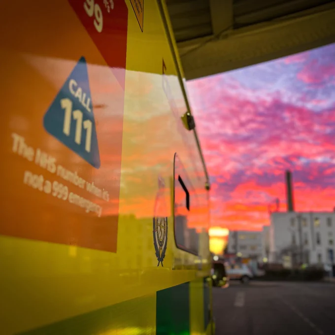 Image shows an ambulance in a car park with a colourful pink and purple sky. The message on the side of the ambulance reads: 'Call 111 - the NHS number when it's not a 999 emergency'.