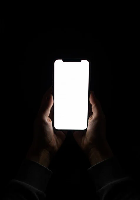 Two hands hold a smartphone with a brightly lit, blank screen in a dark environment. The phone’s screen casts a stark white light, illuminating the hands and creating high contrast against the pitch-black background. The composition focuses on the phone and hands, with the screen as the central point, framed symmetrically from a front perspective. The mood is minimalistic and mysterious, emphasizing the isolation of the device and the darkness surrounding it.