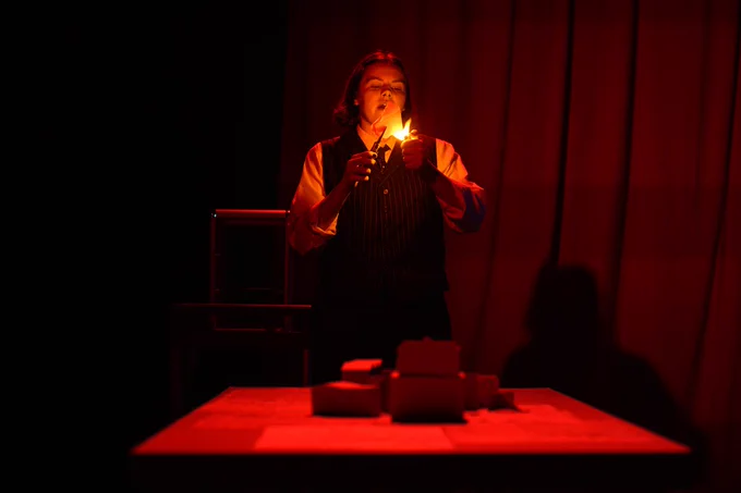A white woman dressed in a white shirt and black pin-striped waistcoat stands behind a table and sets a piece of paper alight. The lighting is a warm orange.