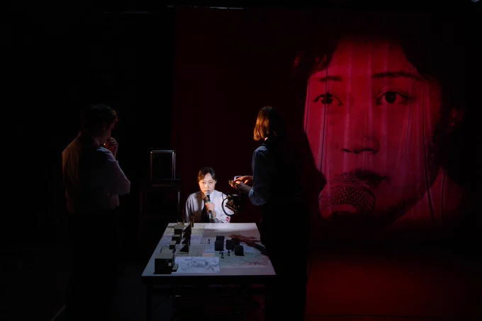 An ESEA woman sits at a white table with models in front of her. Another woman points a camera at her, projecting her face in large scale onto a red curtain behind her.