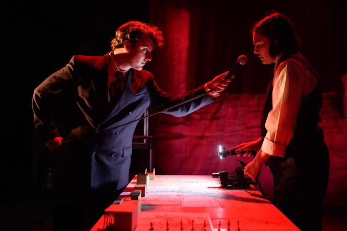 Two people bathed in red light stand on opposite sides of a table. On the left, a white man with curly hair is dressed in a suit. He holds a microphone out to the woman on the other side of the table.