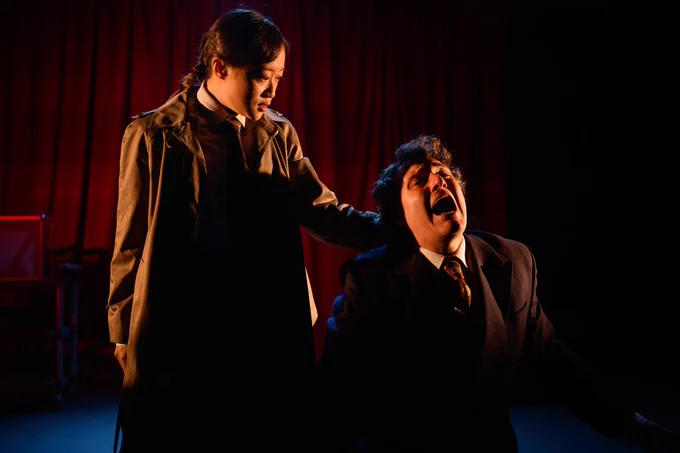 A young woman dressed in a trench coat stands holding a man by the scruff of the neck. He is kneeling on the floor in a submissive position and crying out in pain or desperation.