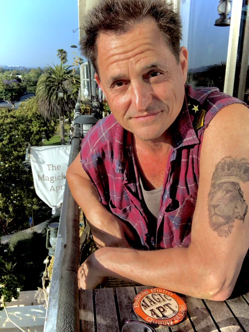 MAGICKER Mike Michael DeLuise sincere sunny  smile in cut off red shirt leaning in w/ Lion arm tat  & The Magic Apt. coaster on Santa Monica view balcony age 55, born Aug 4th, 1969 #MagickerMike MagickerMike.com #TheMagicApt. *since 1999, last century.