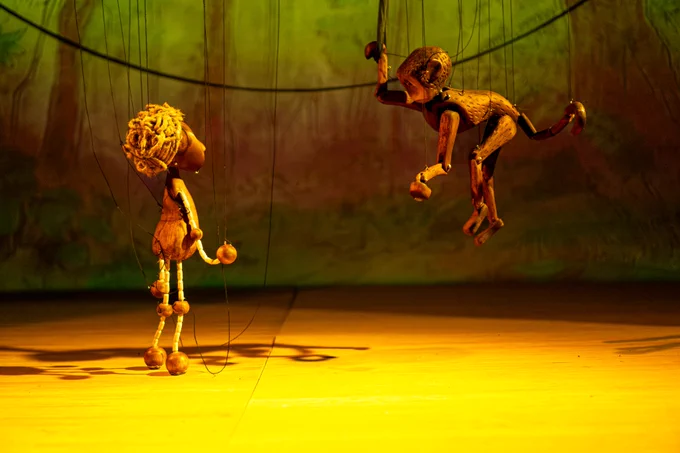 A wooden doll marionette with curly hair is in a forest looking up at a wooden monkey hanging from a vine