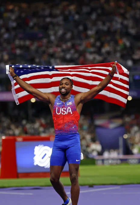 Noah Lyles holding up the American flag as he walks on the track after winning the men's 100m.