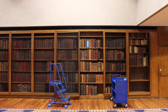 A wall lined with wooden bookshelves, filled with old brown leather-bound books. A set of blue library steps and a blue library trolley are in front of the bookcases. 