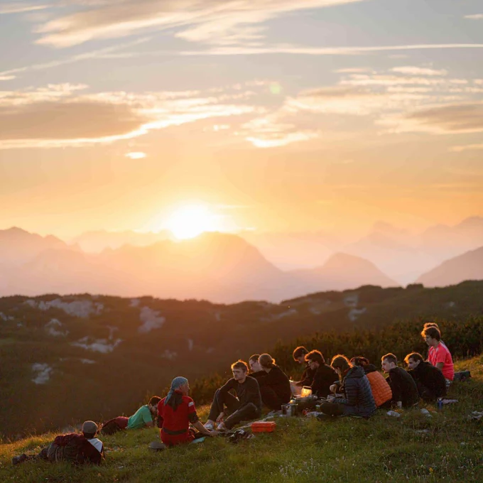 A group of cavers sitting in the grass, enjoying the sunset on the top of the mountain, eating dinner.