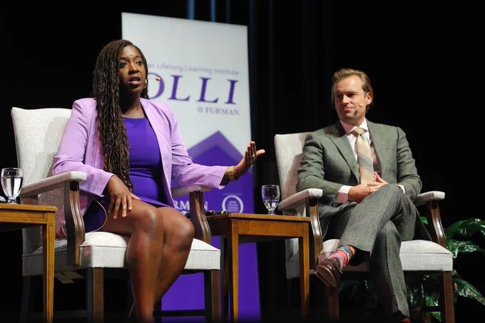A Black woman (left) and a white man (right) wearing mics sit on stage and talk to audience.