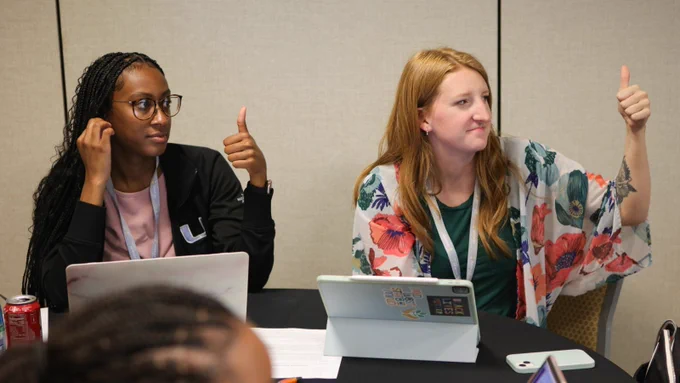 Two women educators sitting beside each other engaged in a professional learning breakout session. They are both shown smiling with thumbs up looking off-camera.
