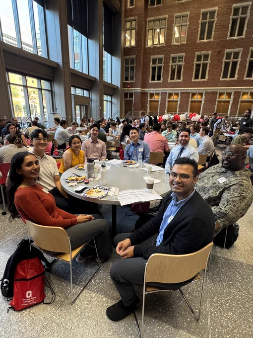 A group of seven people, smiling and seated around a table with plates of food, inside a spacious atrium with large windows and a high ceiling. The background shows many more people sitting and a vibrant, busy atmosphere. One person is wearing a badge on a lanyard. On a red backpack next to one person, there's a visible Ohio State University logo.