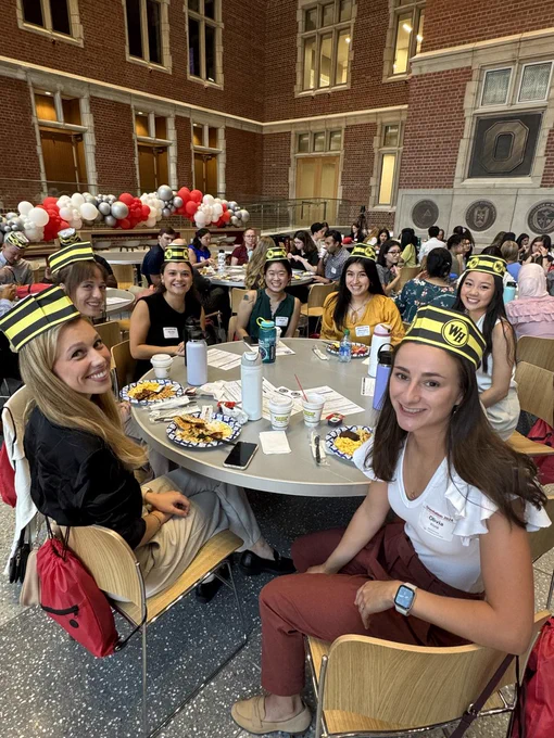 A group of students wearing various hats sit around tables adorned with snacks and balloons in a school hall with brick walls and arched windows.