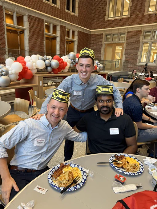 Three people in formal attire, each wearing a distinctive cap, sit at a dining table filled with plates of food in a large hall adorned with red and white balloons. The central individual stands behind the other two who are seated, all smiling at the camera. The setting suggests a celebratory or formal gathering.