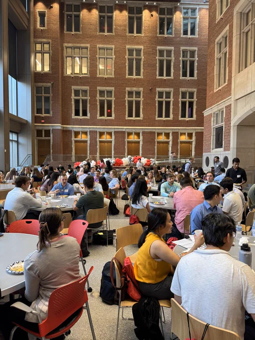 Students dining in a large, atrium-style cafeteria with historical brick building visible through the windows.