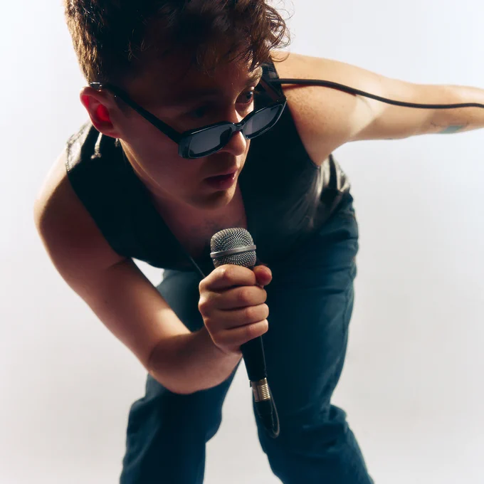 A photo of Jake Roche wearing sunglasses and a black waistcoat and jeans, holding a microphone