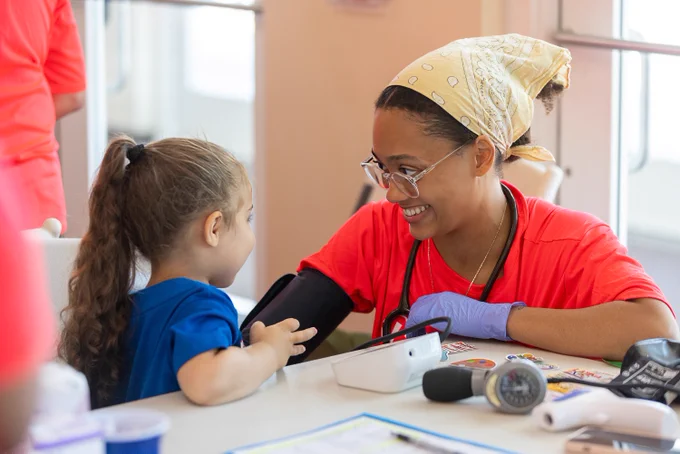 A Rush University student in a red shirt interacts with a young child at a table covered with medical tools and papers. 