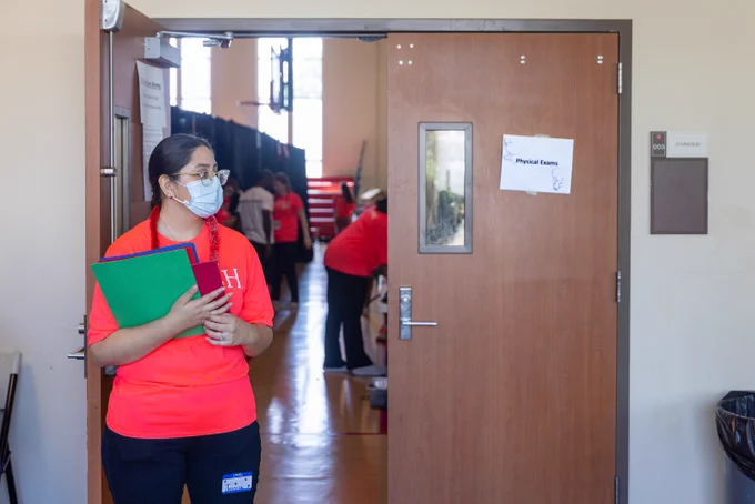 A Rush University student wearing a mask, holding a green folder, walks through a hallway by a room labeled 'Physical Exams,' with several others seen in the background.