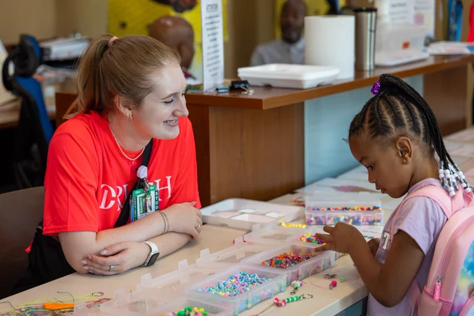 A Rush University student smiles while assisting a young child at a crafting table filled with colorful beads. 
