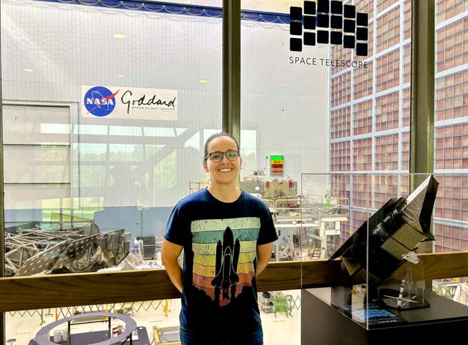 La Dra. Ariadna Farrés Basiana posa sonriente ante un ventanal que da a la cámara limpia del Centro de Vuelo Espacial Goddard de la NASA, donde se está construyendo el telescopio espacial Roman.