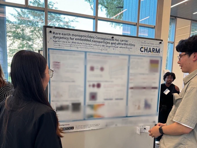 Grad student Ruiqi Hu presenting her research poster to a visitor. Poster is blurred except for the title "Rare-earth monopnictides: Consequences for carrier dynamics for embedded nanoparticles and ultra thin films."