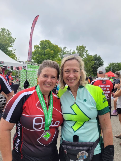 Two participants smiling at the camera after a cycling event, wearing sportive attire and medals. One is in a red jersey with the "9010" logo, and the other in a green jersey with a bold arrow design, against a background with event tents and a crowd.