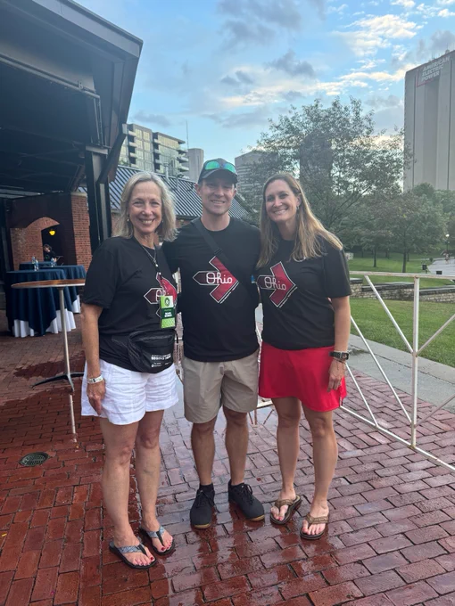 Three people wearing event t-shirts with "Ohio" and Pelotonia logos standing outdoors, smiling at an event with buildings and trees in the background.