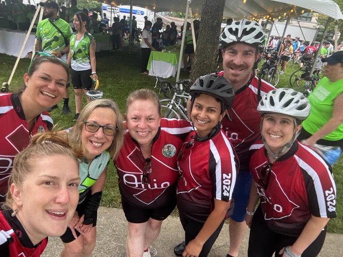 A group of seven cyclists in matching red and white "Ohio" jerseys taking a selfie at an outdoor event with other cyclists and tents in the background.