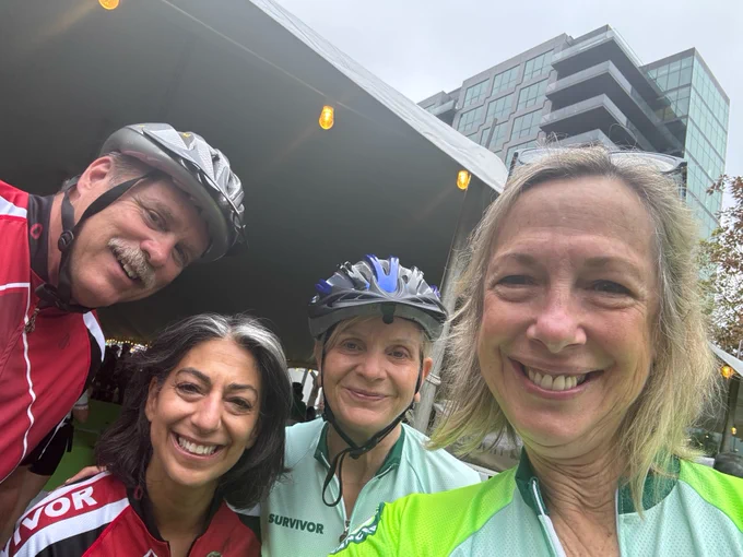 Four adults in bicycle helmets and cycling gear smiling for a selfie, with a modern building in the background. Two are wearing jerseys labeled "Survivor".