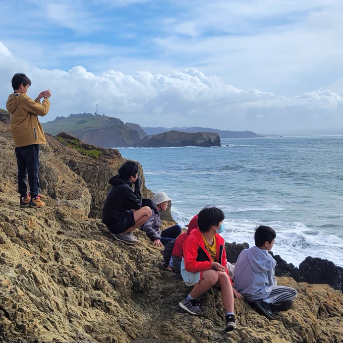 A group of students sits and stands on a rocky shoreline looking out at the waves of the Pacific Ocean.