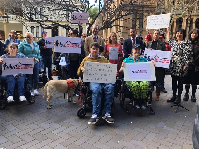 A group of people including campaign spokespeople Julie Charlton and Declan Lee, as well as Alex Greenwich, Jenny Leong and Abigail Boyd from NSW Greens and others gather in front of NSW Parliament holding placards that read End Housing Discrimination Now. 