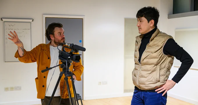 Two individuals are engaged in a video production activity in a studio; one person is operating a camera while the other, wearing a vest, stands by, preparing for a scene.