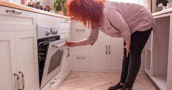 Image of woman wearing a grey top and black jeans is opening an oven door. White cupboards in the background.