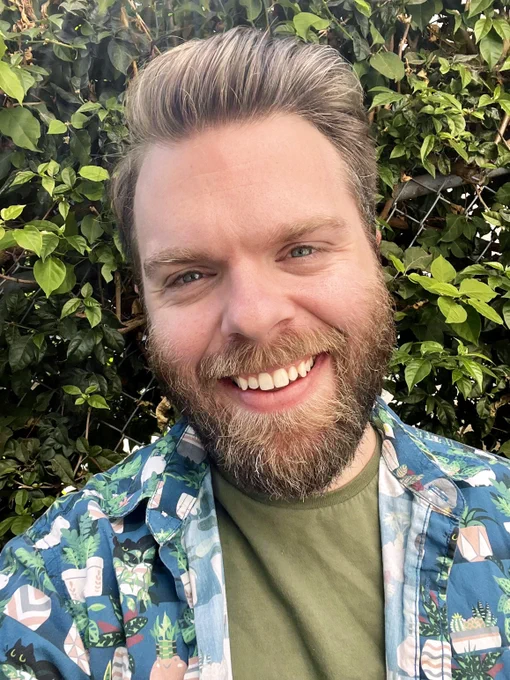 Outdoor selfie of robo smiling in front of a leaf covered chainlink fence. He wears a green t shirt covered by a blue patterned button down, worn open. The pattern on the blue shirt consists of cartoonish black cats hiding among potted plants.