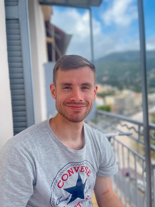 Callum sat on a balcony, with mountains and the sea in the background, in Menton, France, smiling at the camera.