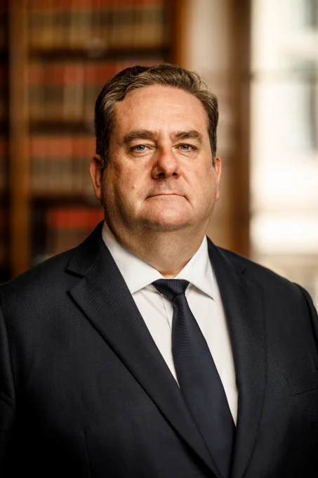 Photograph of Nick Emmerson. Middle aged man with short brown hair, wearing a black suit and blue tie. He is standing in the Law Society library, with a set of bookshelves behind him.