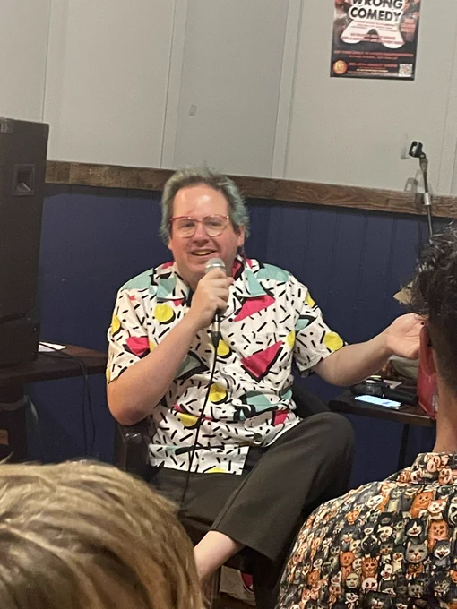 Friz, a non binary person in a loud short sleeved shirt holds a microphone and looks confused