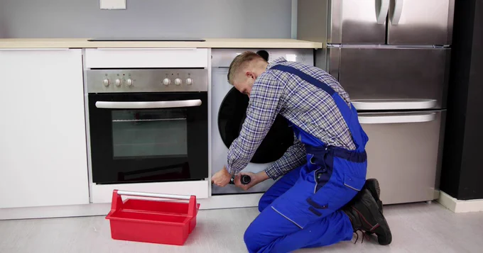 Image of man wearing a blue checked shirt, blue work dungarees, and black boots. The man is fixing a washing machine, holding a screwdriver on the door of a washing machine. There is a red tool box on the floor, white cupboards, a black and grey oven, a silver fridge.