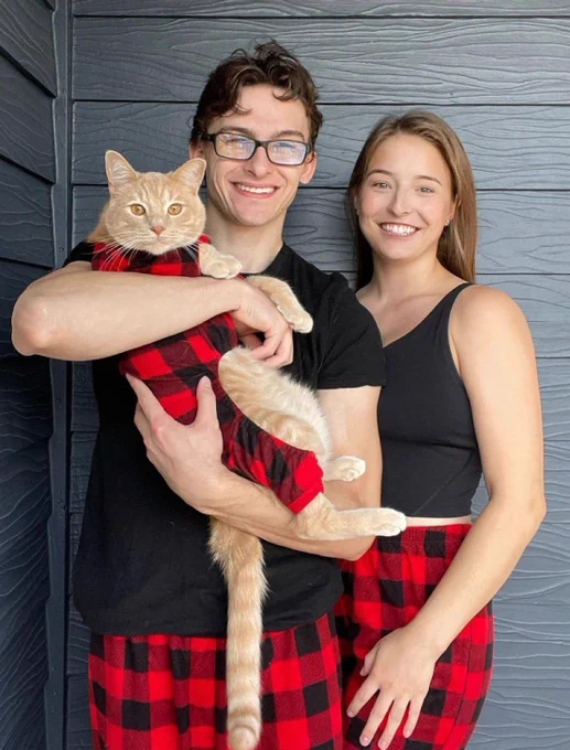 Olympic medalist and pommel horse guy Stephen Nedoroscik holding his orange cat and standing with his girlfriend. All three are wearing matching red buffalo plaid pajamas