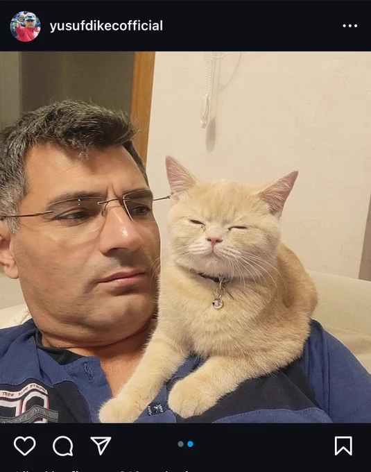 Turkish Olympian slash casual pistol shooter slash like bad guy cop sorry Yusuf Dicek sitting on a sofa with his sleepy orange cat who is probably too dumb to have done anything evil on his shoulder