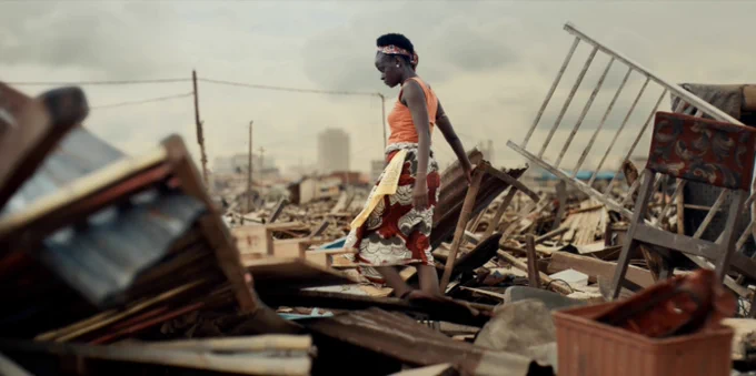 A film still from The Legend of the Vagabond Queen of Lagos. A woman walks through an enormous lot of broken furniture. It looks like it could be the aftermath of a natural disaster. She is wearing an orange tank top and a red patterned skirt, and she has a red patterned headband on. There are telephone poles visible behind her, and in the distance, outlines of tall buildings.