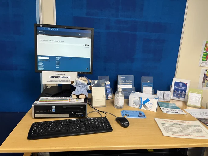 A computer set up at a desk, with Library Search on the screen. A toy monkey in hospital scrubs sits next to it. 