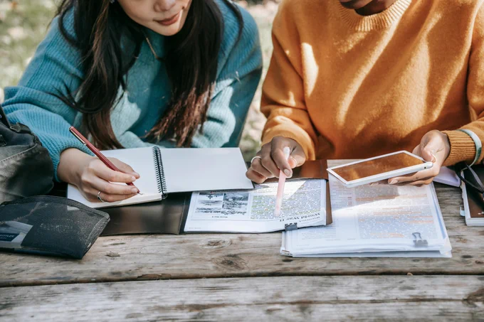 Two people sitting at a table with a notebook and tablet, closely examining a piece of text in front of them.