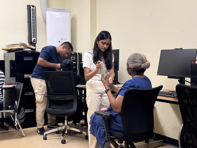 In a computer lab, two older adults sit and practice using a tablet. An instructor and a volunteer are each helping one of them.