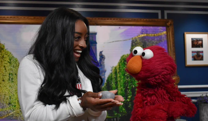 Simone Biles, smiling, holding Rocco with both hands and looking at Elmo