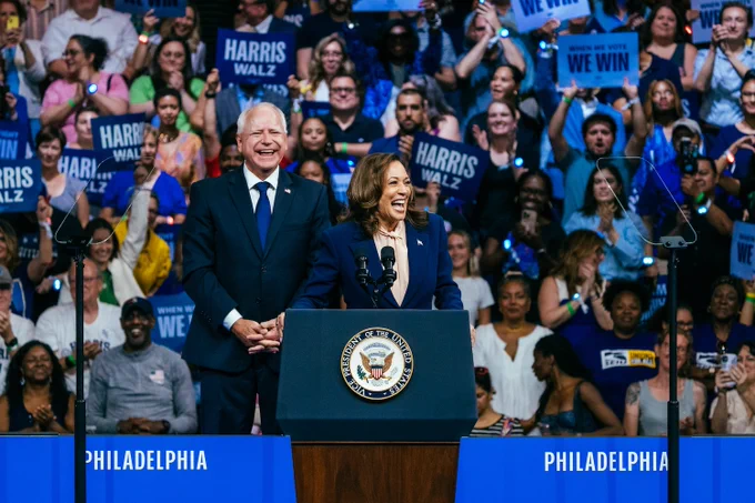Vice President Kamala Harris stands in front of Governor Tim Walz at a podium in front of a crowd holding blue Harris Walz signs.